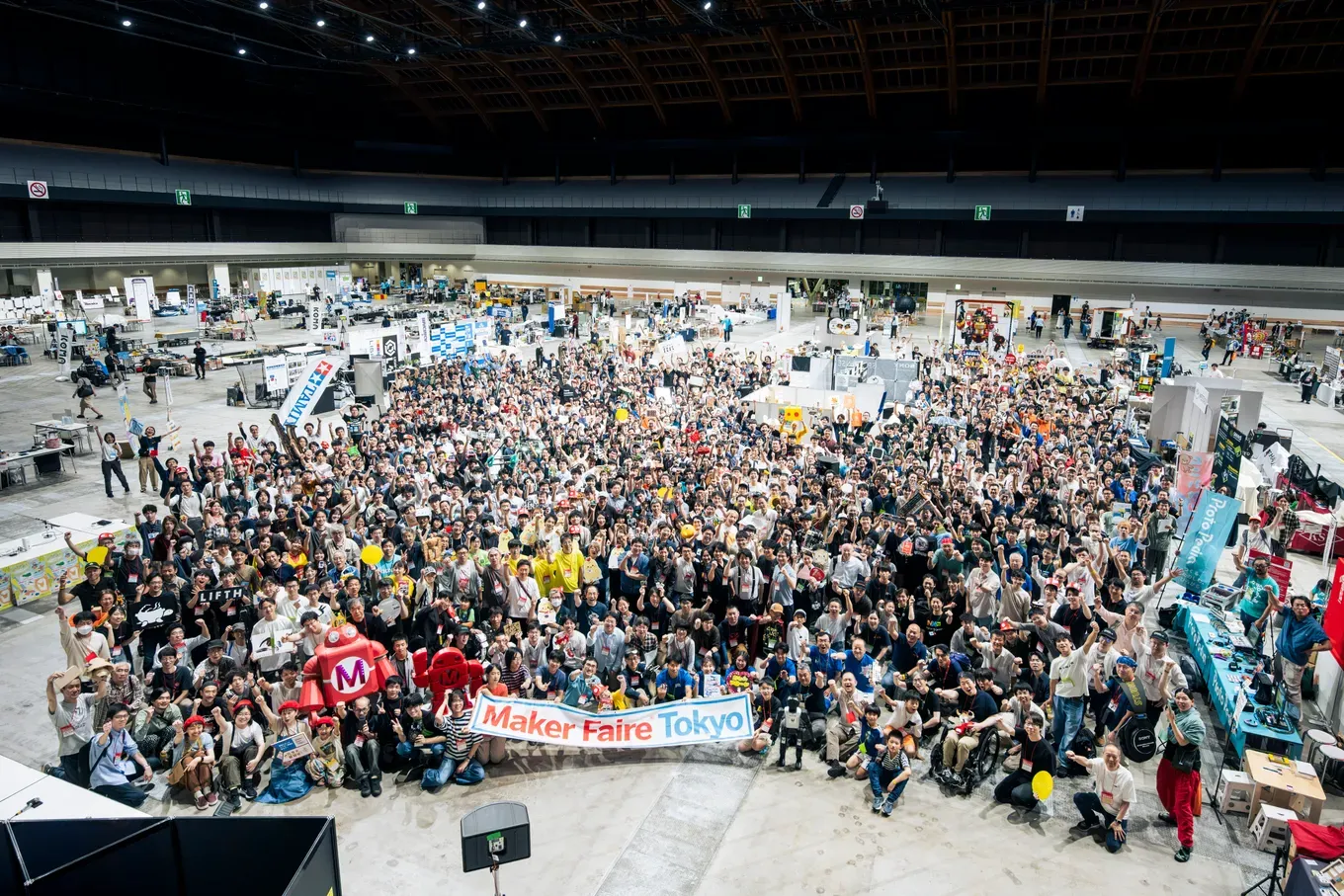 Maker Faire Tokyo 2026 の会場で、出展者と来場者が「Maker Faire Tokyo」の横断幕を囲んで手を上げている集合写真。上から見下ろした構図